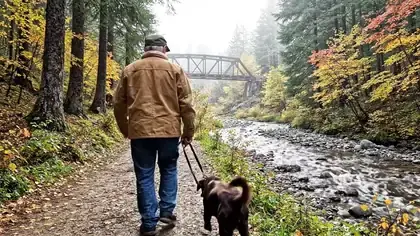 A man in a work jacket walking a brown dog along a creek trail toward an old iron bridge on a misty morning