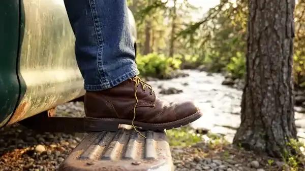 Weathered hands tying the laces of a work boot on the running board of a pickup beside a creek